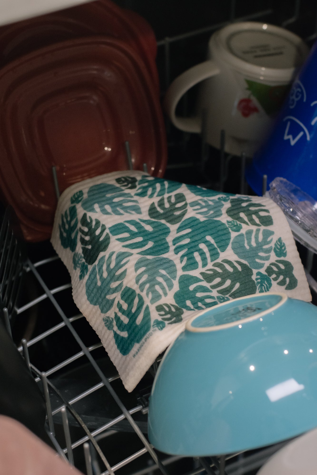 Dish towel with green leaf pattern hanging in a dishwasher with mugs and bowls.