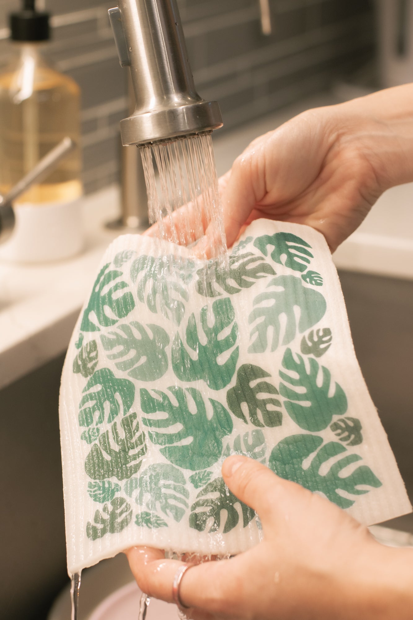Person washing a reusable cloth with green leaf pattern under running water in a kitchen sink.