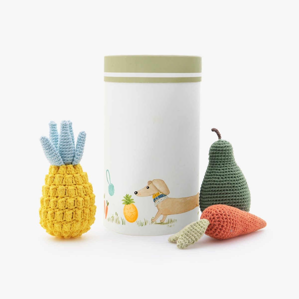 White container with a dachshund design for the Organic Cotton Veggie Dog Toy Bundle – Eco-Friendly Squeaky Carrot, Pear & Pineapple Set - accompanied by the crocheted dog toys - yellow pineapple, green pear, and orange carrot - on a white background.