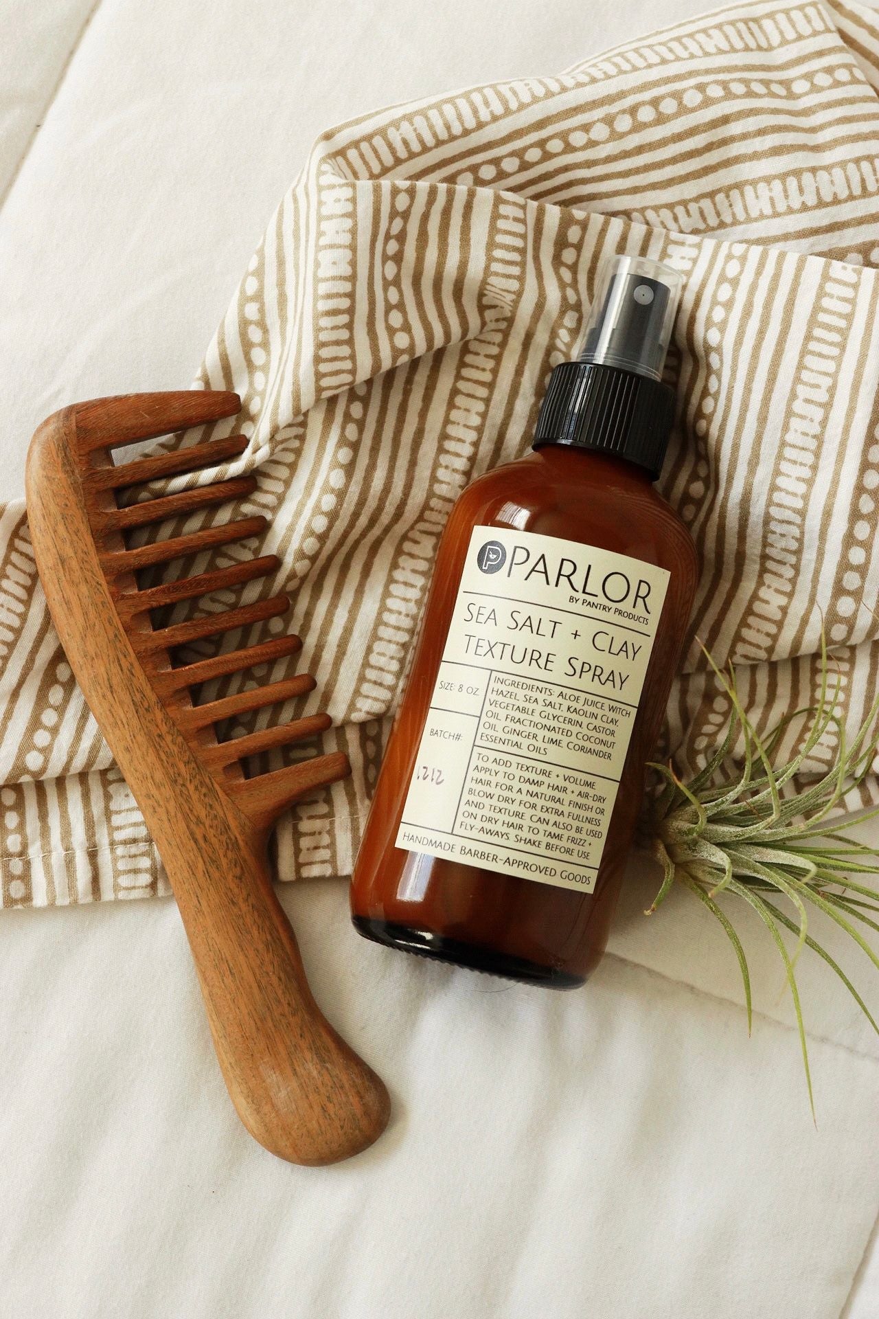 Amber glass bottle of Parlor Sea Salt + Clay Texture Natural Hair Styling Spray with a wooden comb and plant on a textured fabric background