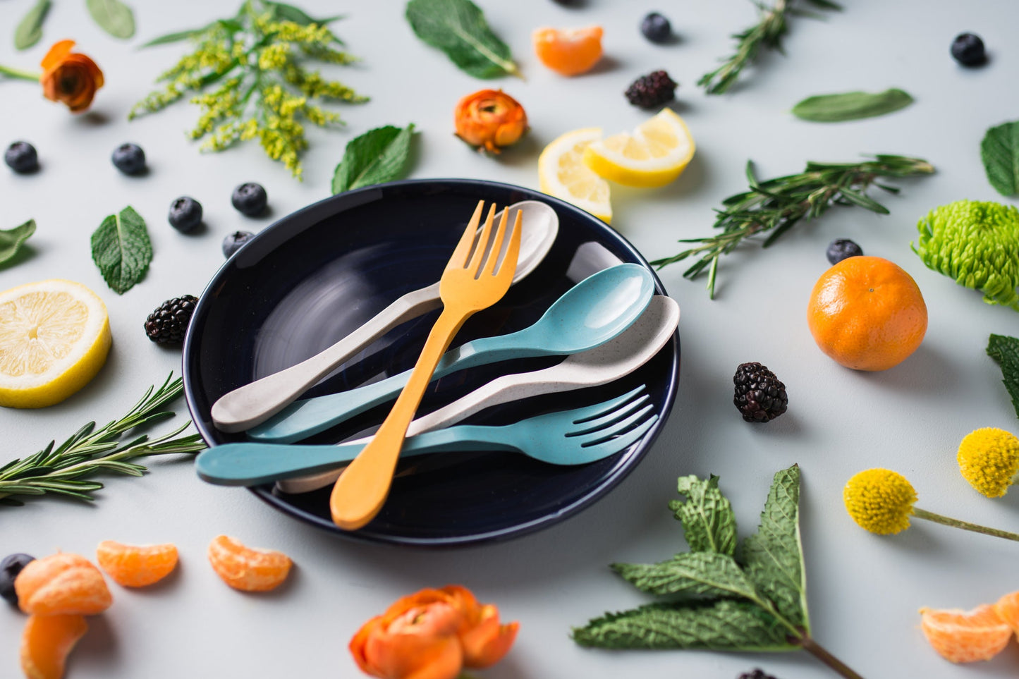 Set of 8 colorful compostable forks and spoons on a plate with fruits and vegetables arranged around it. Plastic-free bamboo cutlery.