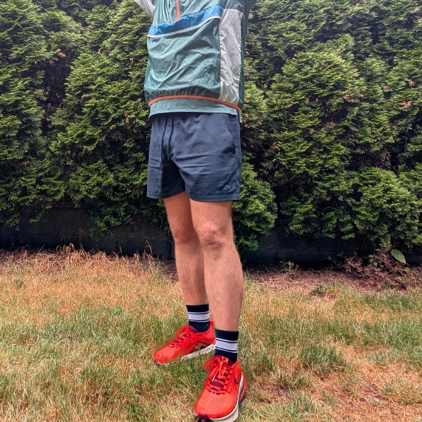 Man wearing a green jacket, Athletic 5 Inch Recycled Shorts by Rainier Watch, and red running shoes standing in a grassy area with green bushes in the background.