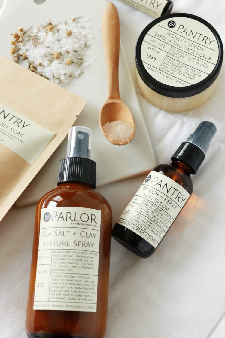 Two bottles of products by Pantry: 'Parlor Sea Salt + Clay Texture Spray' and 'Pantry Refresh & Repair Skin Serum' on a white surface with wooden spoon and salt.