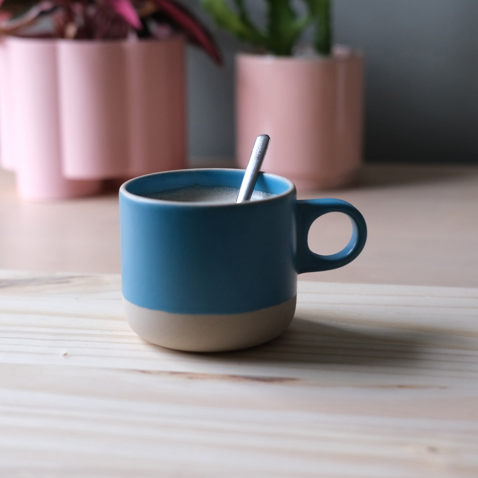 Handmade ceramic blue mug with natural base on a blonde wooden surface, surrounded by pink plant pots. Made in the USA