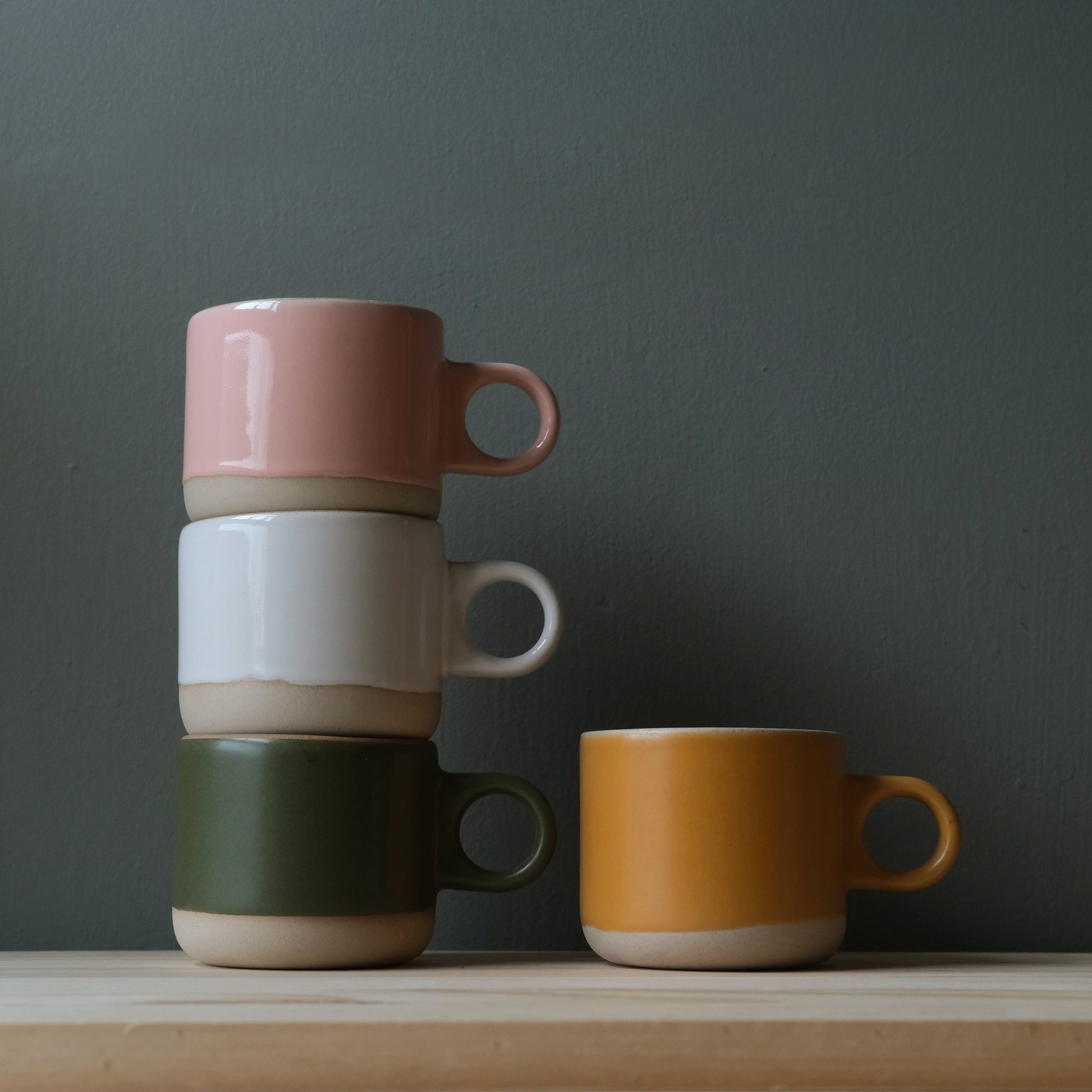 Four colorful handmade ceramic mugs stacked and placed on a wooden surface with a dark gray background. Pink, White, Green and Gold. Made in the USA