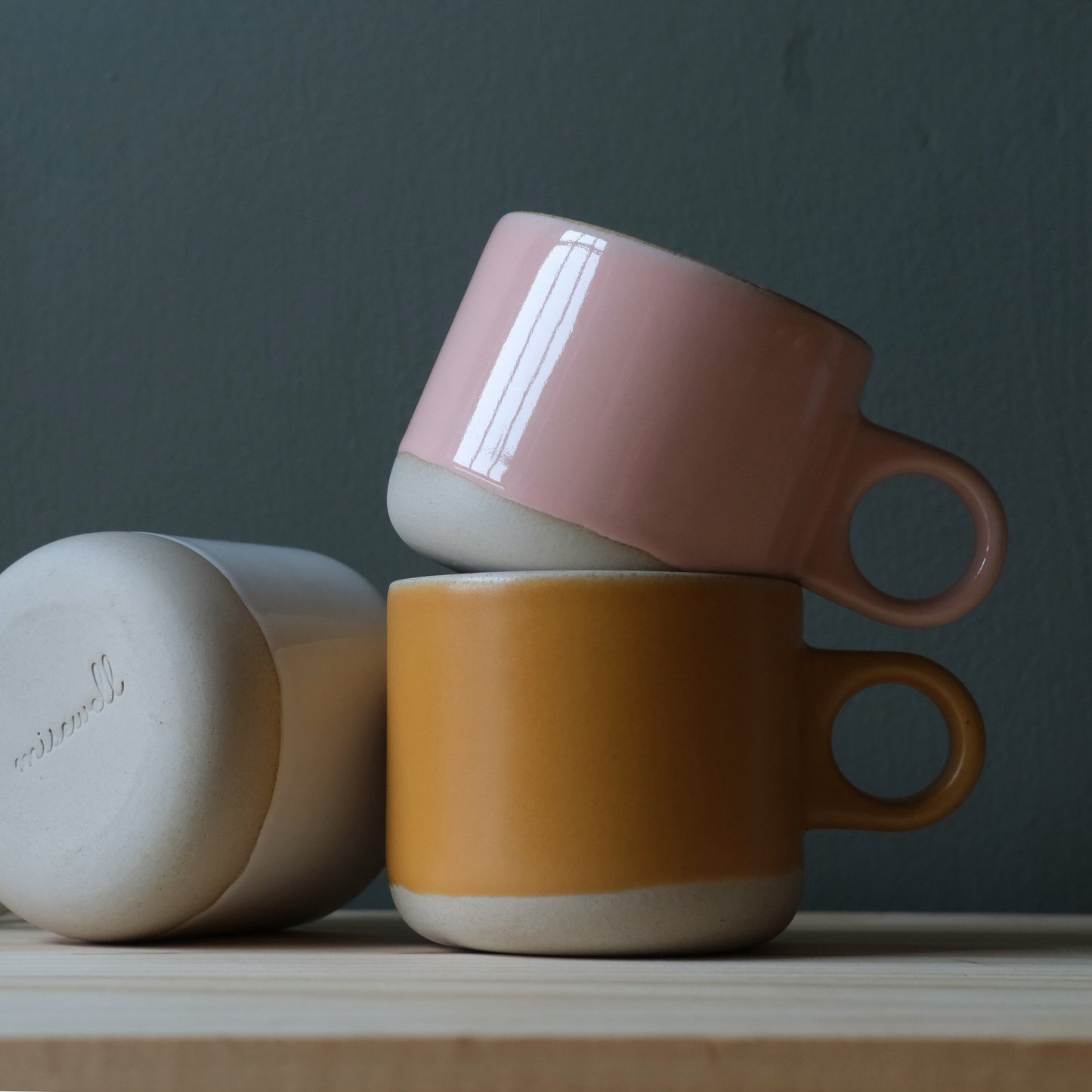 Three handmade ceramic mugs in pink, orange, and beige on a wooden surface with a dark background. Made in the USA