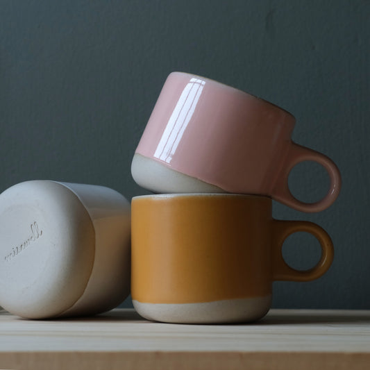 Three handmade ceramic mugs in pink, orange, and beige on a wooden surface with a dark background. Made in the USA