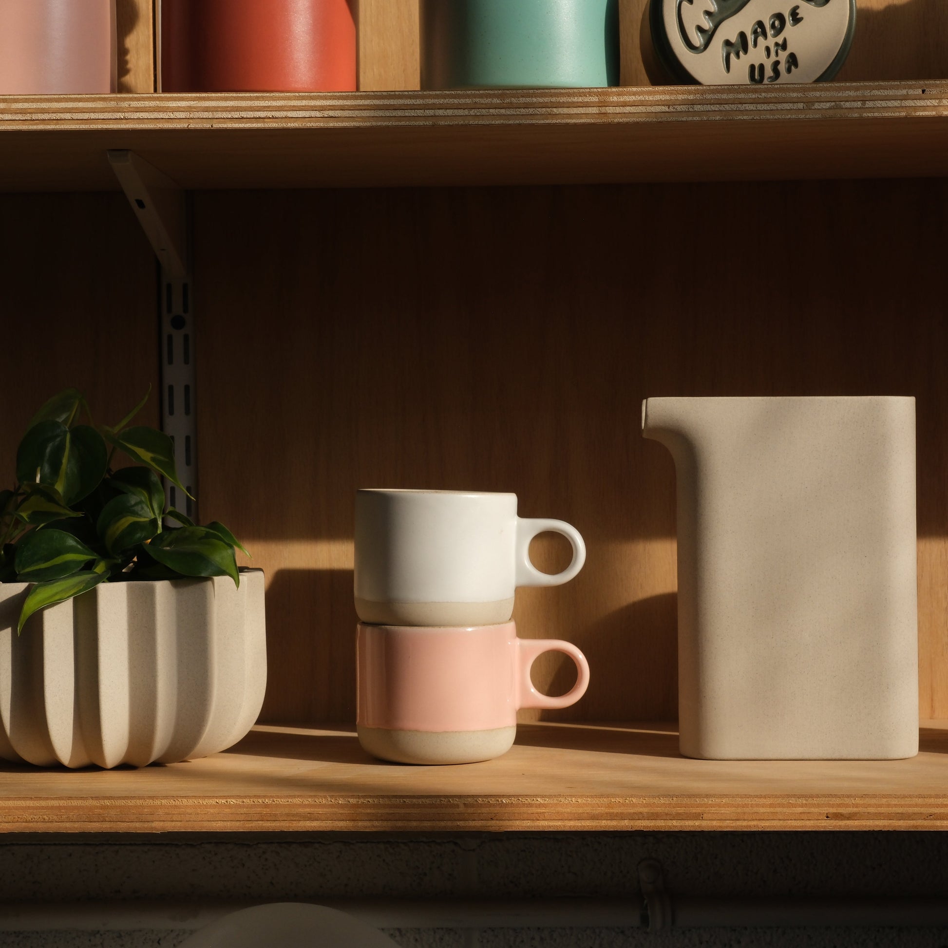 White handmade ceramic mug and pink handmade ceramic mug on a wooden shelf with a pitcher and plant. Made in the USA