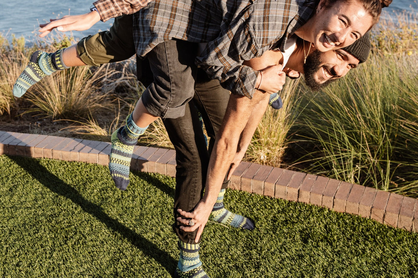 Two people playfully interacting on a grassy area with a scenic background in mismatched Solmate Lemongrass Crew Socks
