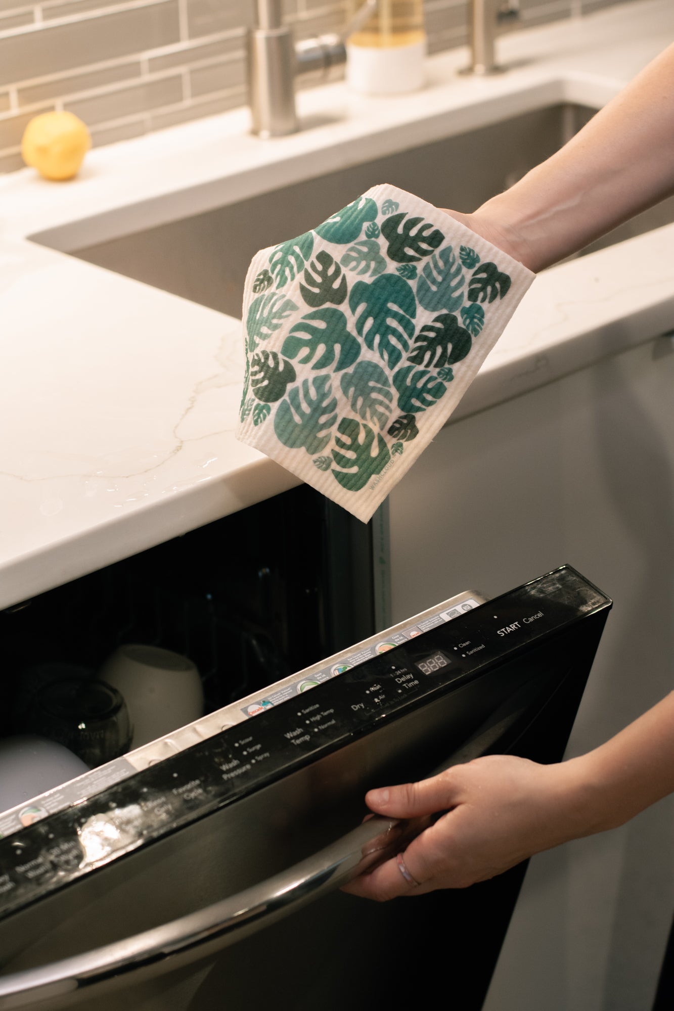Person opening a dishwasher with a green leaf-patterned cloth.