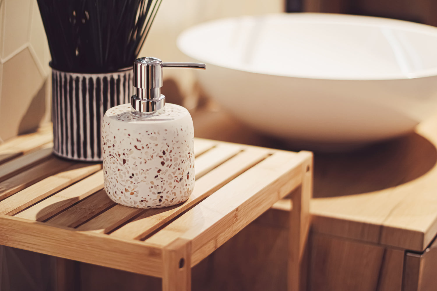 A speckled white soap dispenser with a silver pump bottle, placed on a light wood slatted stool in a warm, modern bathroom setting with a white vessel sink in the background.