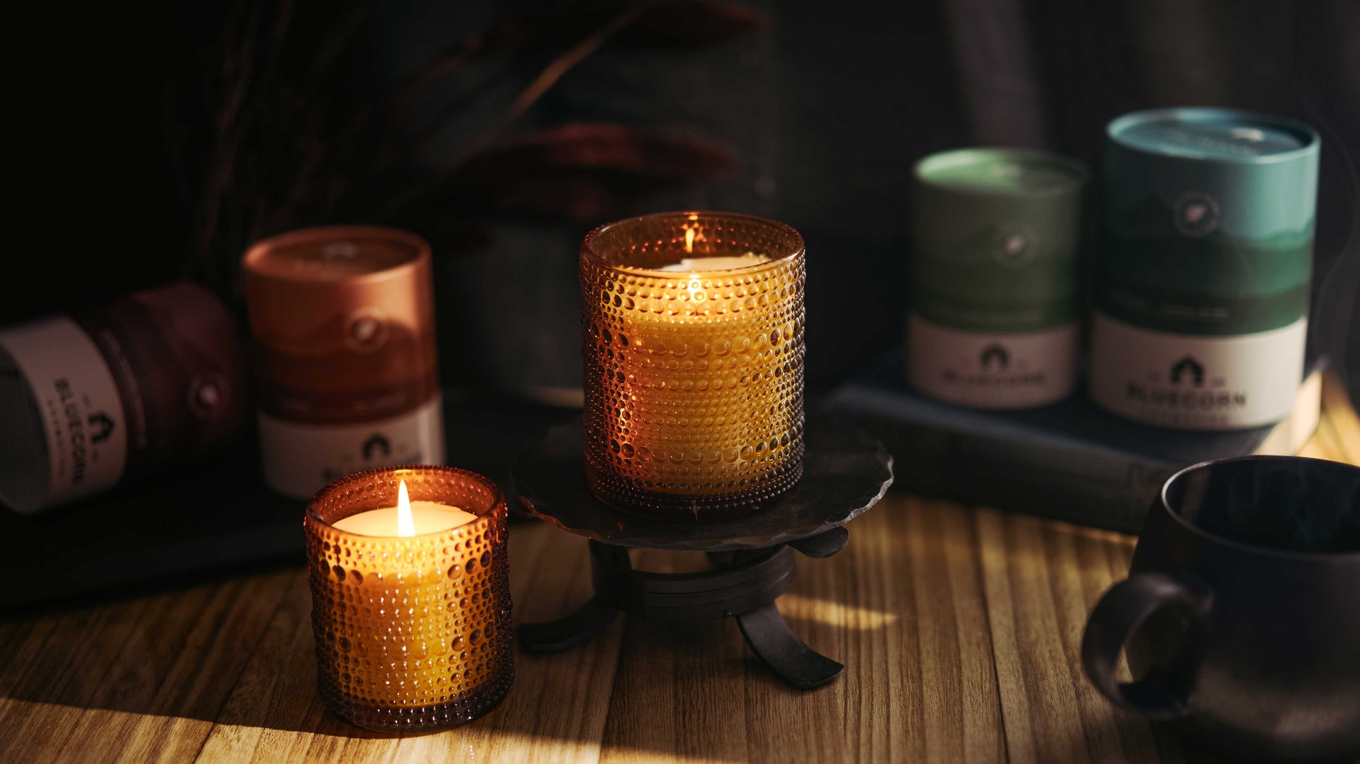 Candlelit scene with Bluecorn Aromatica Winter Scented Candles in textured glass on a wooden surface, surrounded by paper-based packaging and a mug.